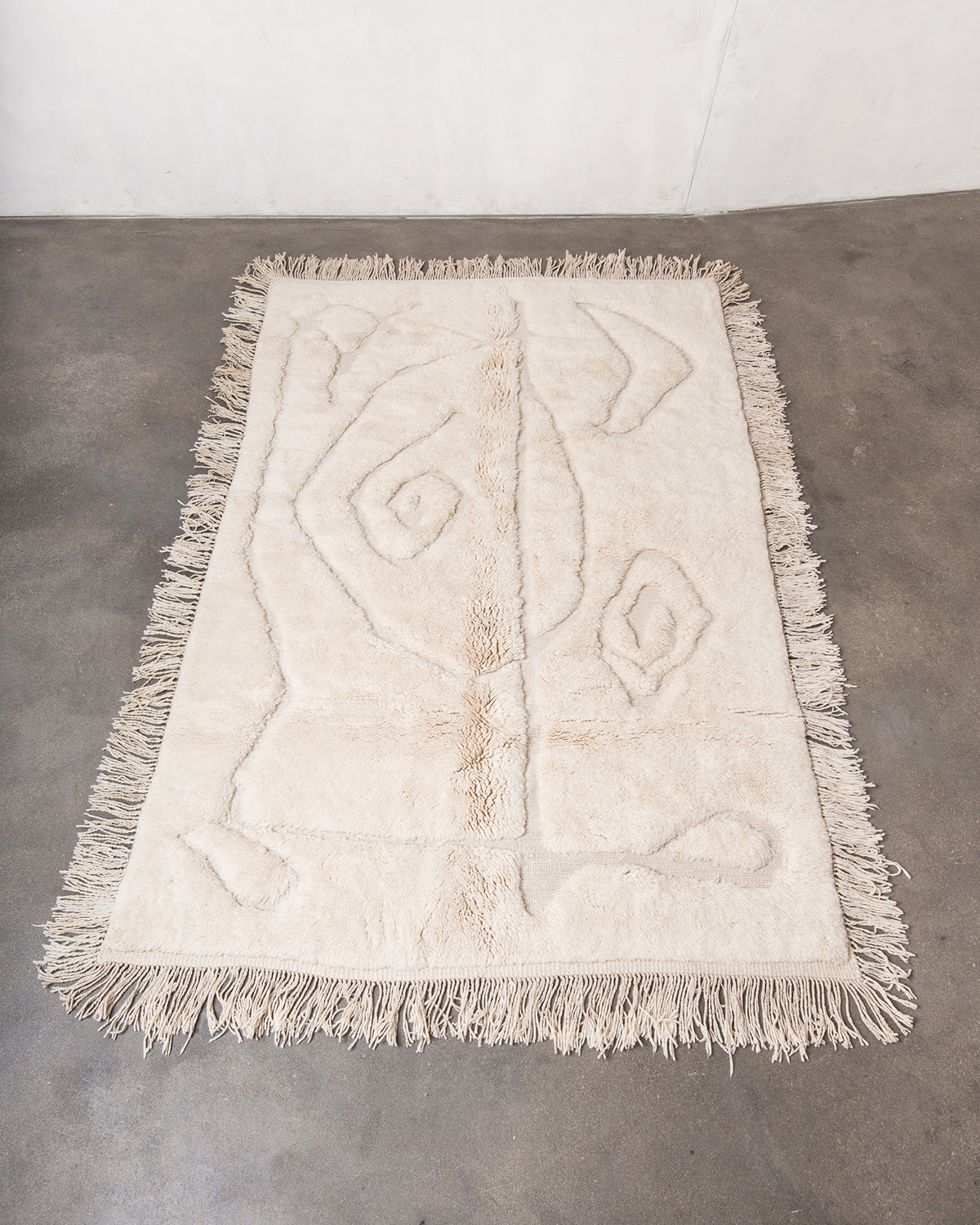 Berberlin Beni M'rirt rug with abstract swirly design by Malaika Raiss. Made of cream sheep's wool with natural wool tassels on all four sides, laying on concrete studio floor.